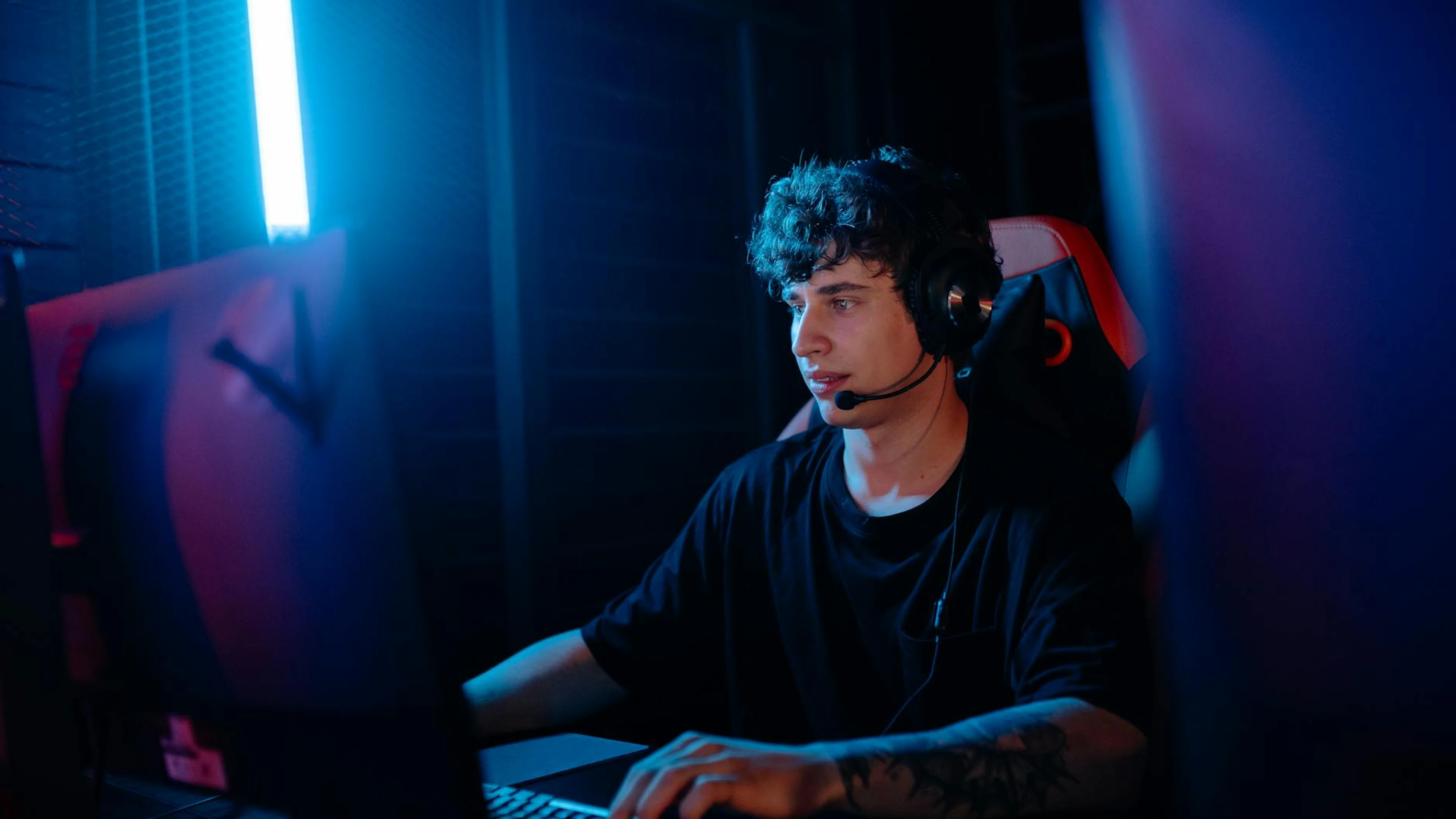 A young man intensely gaming with a headset at a computer desk, illuminated by b - free 4K Ultra HD gaming wallpaper for desktop
