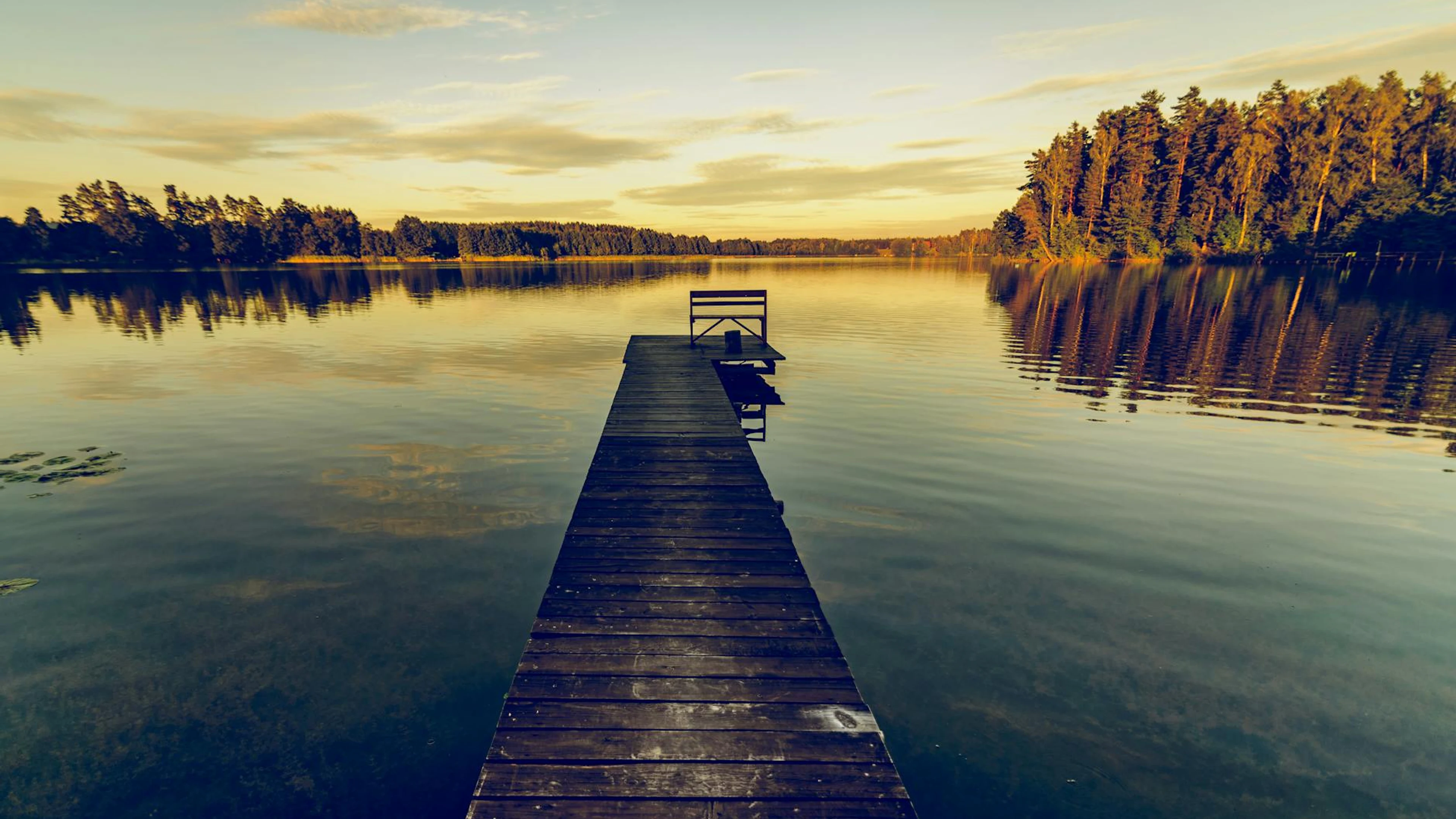 Serene landscape of a wooden jetty over a calm lake at sunset, reflecting trees - free 4K Ultra HD lake wallpaper for desktop