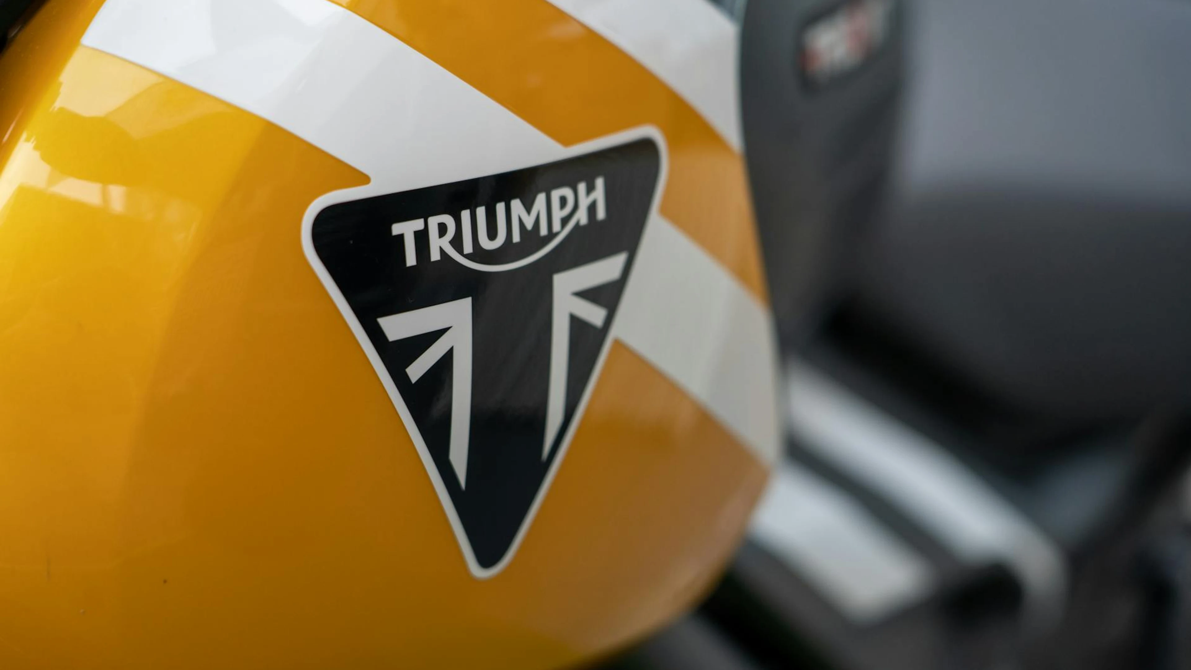 Detailed view of a yellow Triumph motorcycle fuel tank showcasing its logo. - free 4K Ultra HD motorcycle wallpaper for desktop