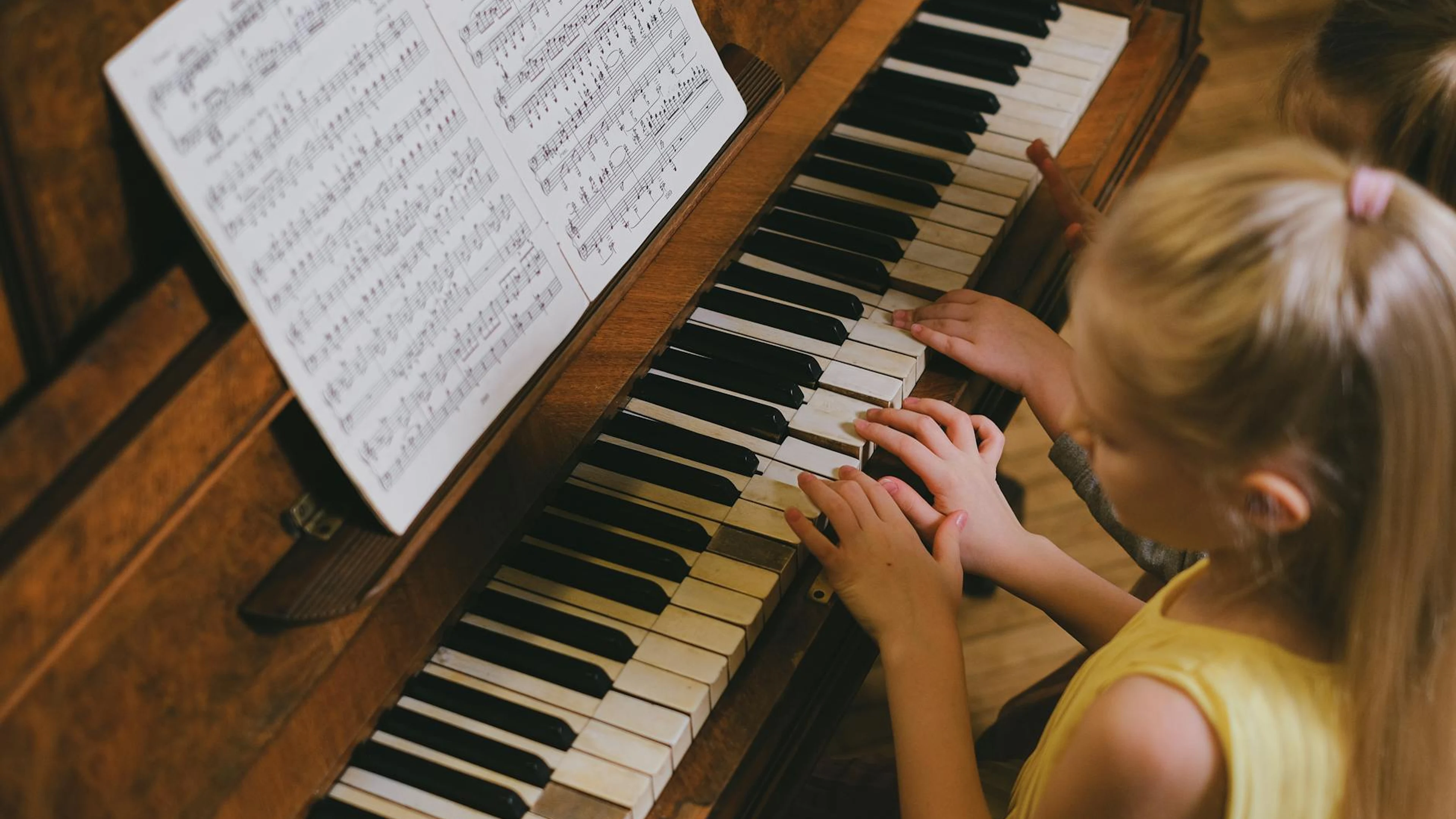 A young girl with blond hair playing piano, following music sheets indoors. - free 4K Ultra HD music wallpaper for desktop