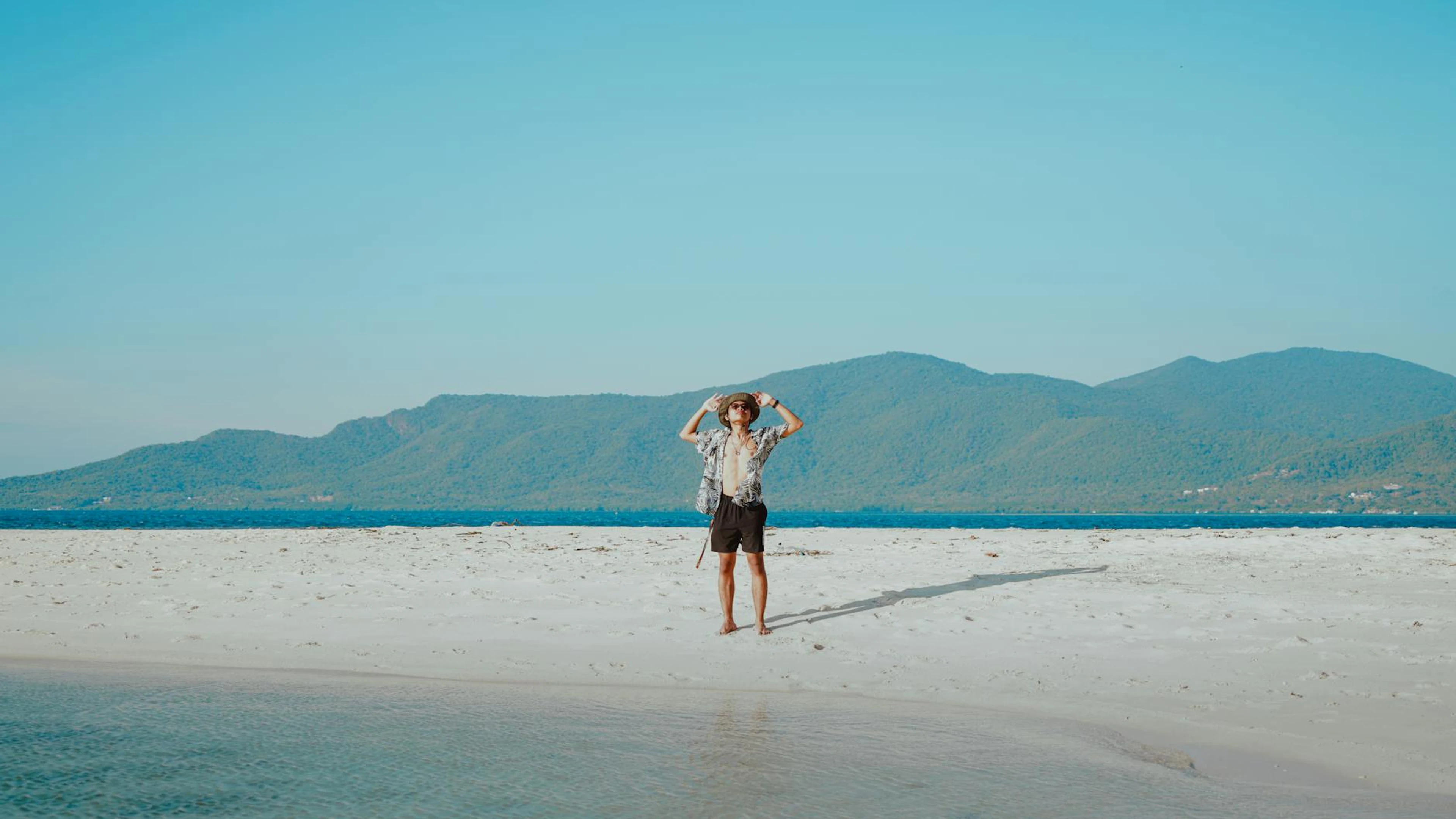 Young man enjoying a sunny day on a serene tropical beach with mountain views. - free 4K Ultra HD nature wallpaper for desktop